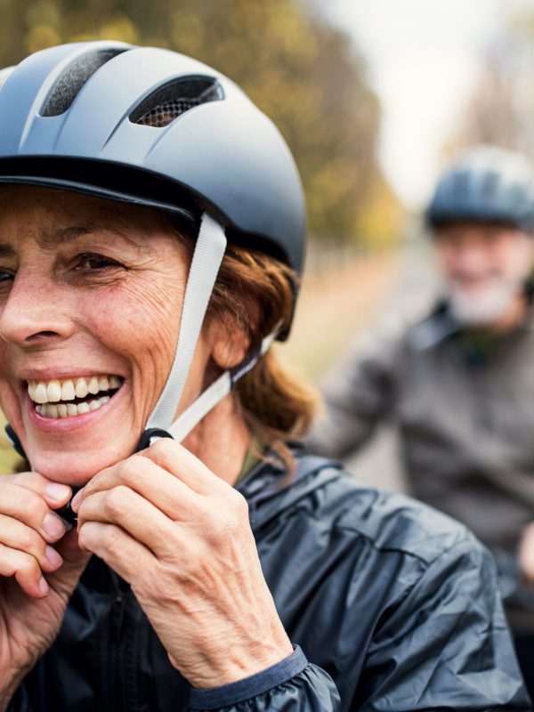 An active senior couple with helmets and electrobikes standing outdoors on a road in nature, putting on a helmet.