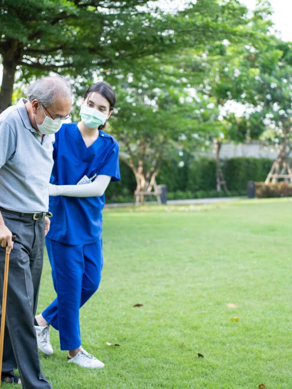 Asian young woman nurse taking care and support disabled senior elderly man on wheelchair at backyard. Caregiver doctor and older mature handicapped patient wear mask to prevent from Covid-19 pandemic